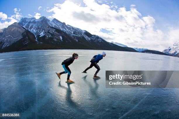 a man leads a woman on a winter speed skating adventure on lake minnewanka in banff national park, alberta, canada. - frozen lake stock pictures, royalty-free photos & images