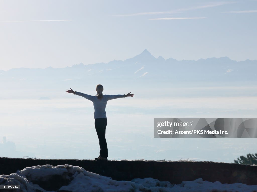 Woman stands on wall, arms outstretched to mtns