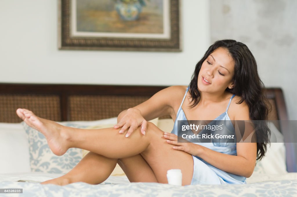 Hispanic woman rubbing lotion on legs in bedroom