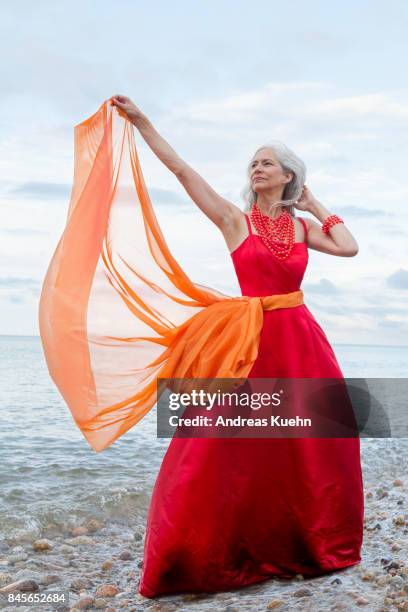 woman with long, grey hair wearing a red evening dress on a rocky the beach while standing in the water. - woman long dress beach stock pictures, royalty-free photos & images