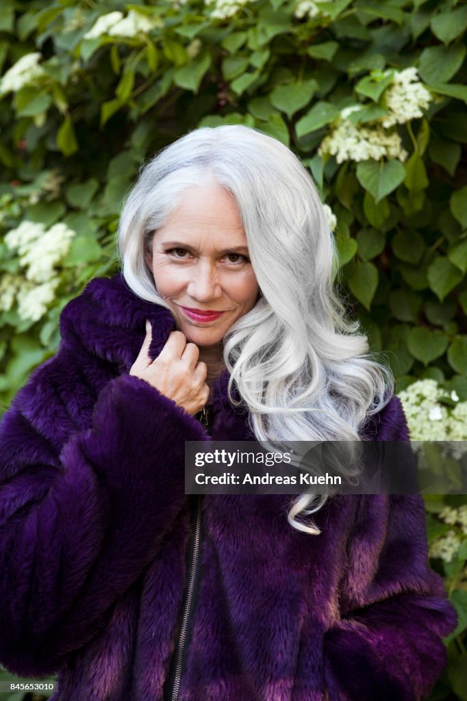 Beautiful woman in a purple faux fur jacket with long, silvery, grey hair and a soft smile standing in front of white flowering ivy, close up.