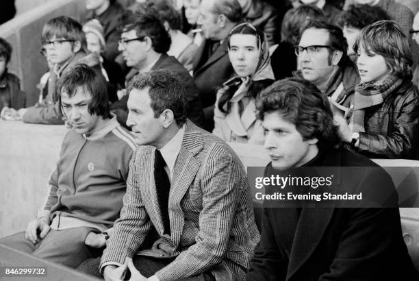 English football manager of Queens Park Rangers Dave Sexton sits on a bench, while following a game against Chelsea FC, with some members of his...