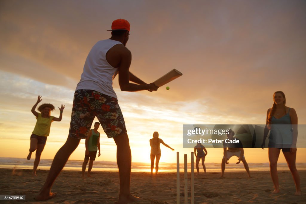 Group of young people playing cricket on beach at sunset