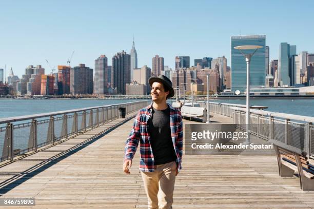 young man in hat walking and laughing against new york city skyline - ragazzo new york foto e immagini stock
