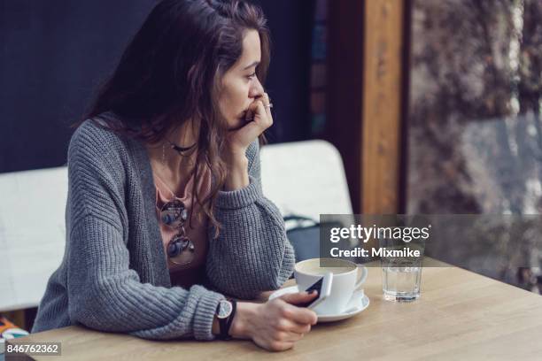 mujer sentada sola, con café y mensajes de texto en su teléfono móvil - pouting fotografías e imágenes de stock