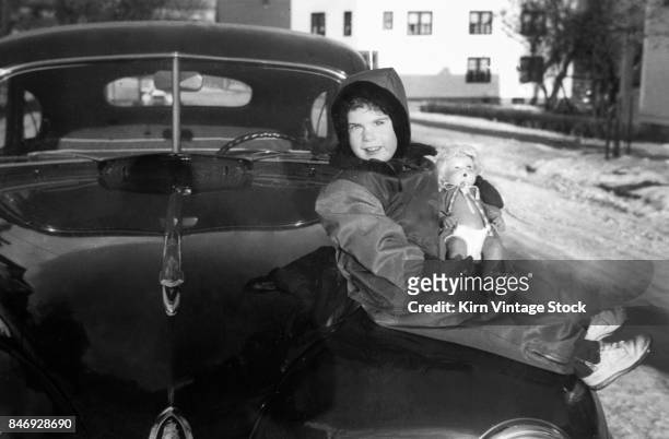 Five year old girl holds her doll and sits on the hood of the family car in the winter.