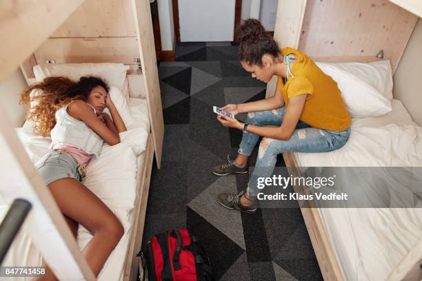 young woman checking phone in bunk bed, roommate sleeping in the other bed - hostel people stock pictures, royalty-free photos & images