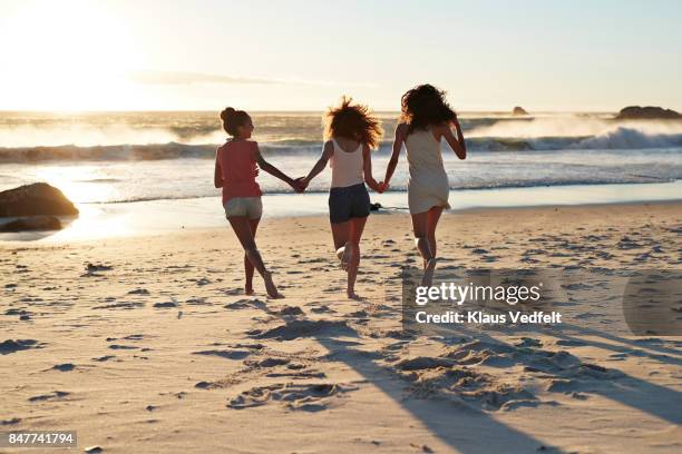 three young women running towards the sea, hand in hand - girl beach sunset stock-fotos und bilder