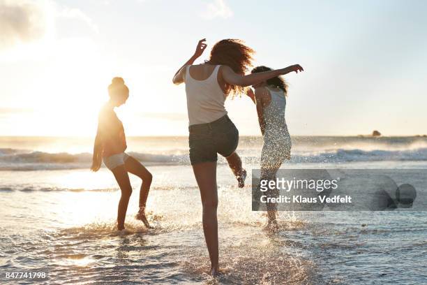 three young women kicking water and laughing on the beach - attività del fine settimana foto e immagini stock