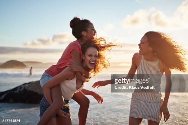 young women piggybacking on sandy beach - strand stock-fotos und bilder