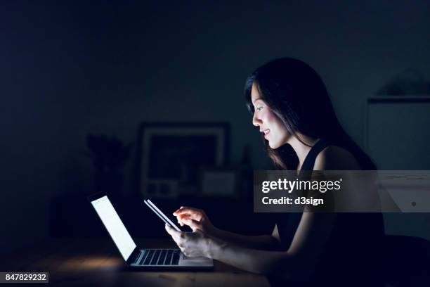 busy young woman text messaging on smartphone while working on laptop till late in home office - working late imagens e fotografias de stock