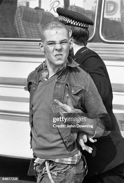 Skinhead is escorted away by a police officer during a bank holiday weekend in Southend, 7th April 1980. The words 'Made in London' and a cross are...