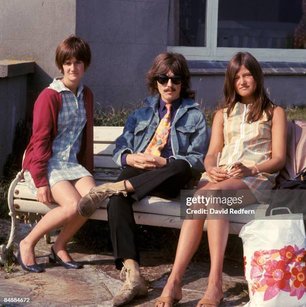 George Harrison of English rock and pop group The Beatles, wearing sunglasses and a denim jacket, sits on a bench with two young women during filming...