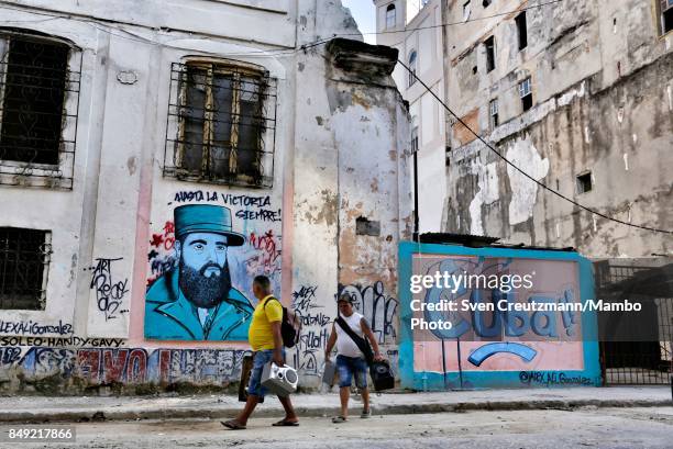 Two Cubans carry audio equipment as they pass by a graffiti that depicts late Cuban Revolution leader Fidel Castro and reads Hasta la Victoria...