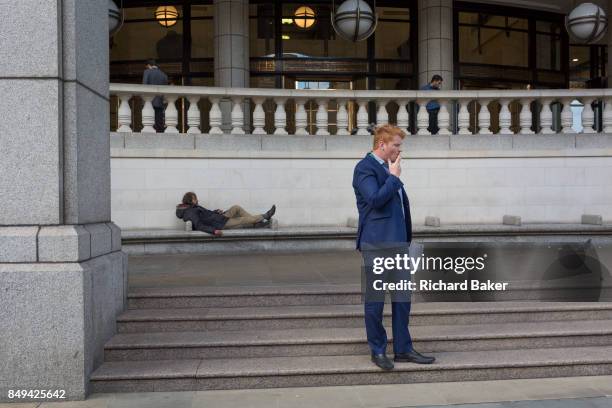 Homeless man lies on City seating as a businessmen smokes, on 14th September 2017, in the City of London, England.