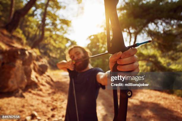 een jonge man bereidt om te schieten met de boog in het bos - archery feather stockfoto's en -beelden