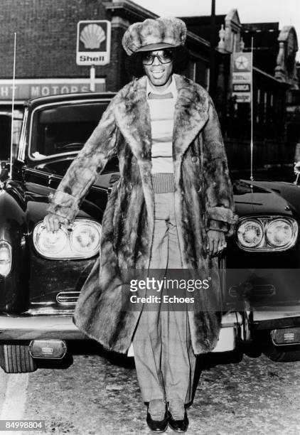 Photo of Johnny Guitar WATSON; Posed full length portrait of Johnny 'Guitar' Watson, standing in front of a car, wearing fur hat and long fur coat...