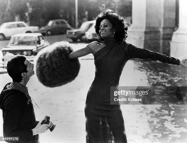 Photo of Diana ROSS; During filming of Mahogany. Posed outide, arms stretched out wearing furry pompom on right arm, photographer smiling looking at...