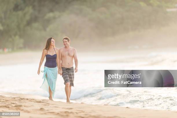 caucasian couple walk hand in hand along a warm hawaii beach - sarong stock pictures, royalty-free photos & images
