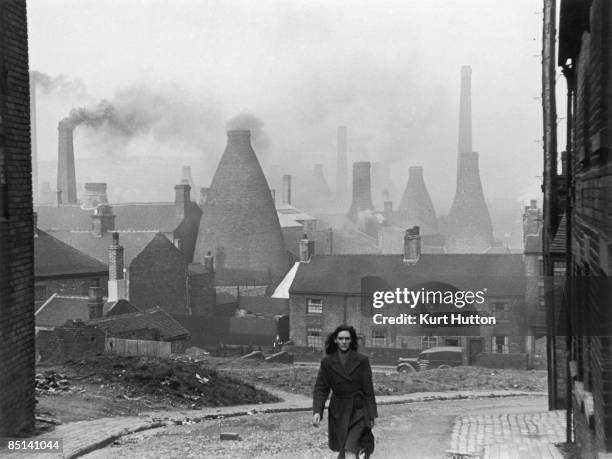 Woman walking up a street in Stoke-on-Trent, Staffordshire, with smoking bottle kilns belonging to potteries visible in the background, 2nd March...