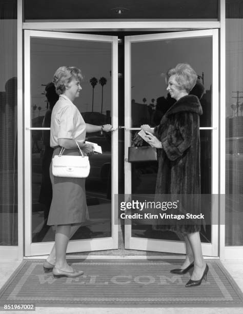 Two well dressed women at bank entrance politely open the door for each other.