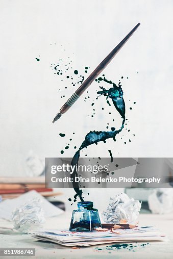 Spilled ink flying above inkwell in a spiraling splash with tiny drops and flying pen on a light background. Still life with writer workplace. Creative writing concept. Spilled ink flying above inkwell in a spiraling splash with tiny drops and flying pen on a light background. Still life with writer workplace. Creative writing concept.