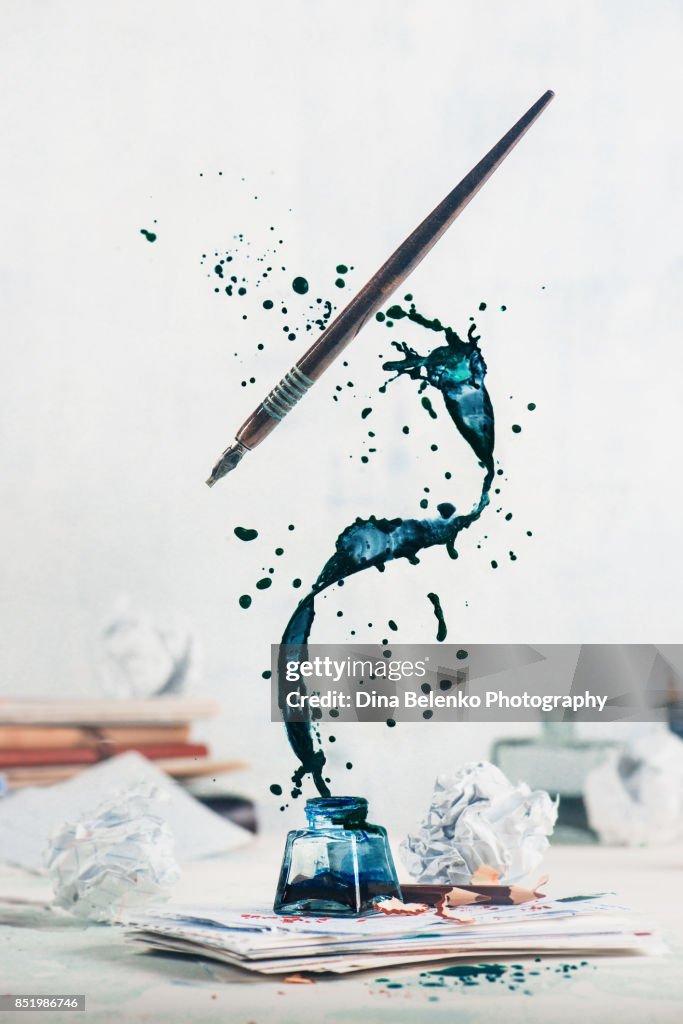 Spilled ink flying above inkwell in a spiraling splash with tiny drops and flying pen on a light background. Still life with writer workplace. Creative writing concept.