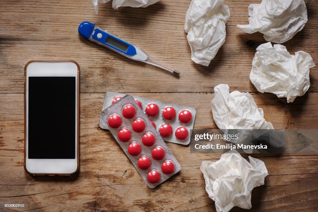 Thermometer, medicine, tissue, mobile phone and a cup of tea on wooden background