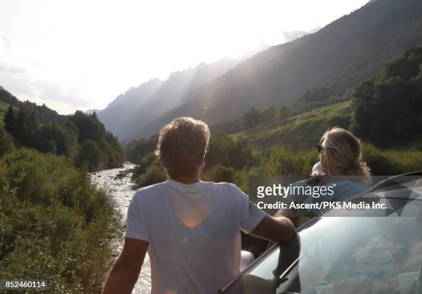 mature couple relax beside car, looks off to mountains, river - car top view photos et images de collection