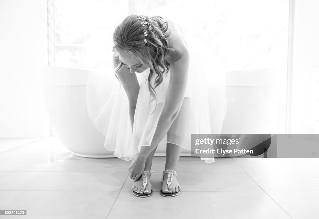 A bride is leaning over to buckle her casual shoes as the finishing touch to getting dressed for her wedding. She sits on a bathtub in front of a large window.