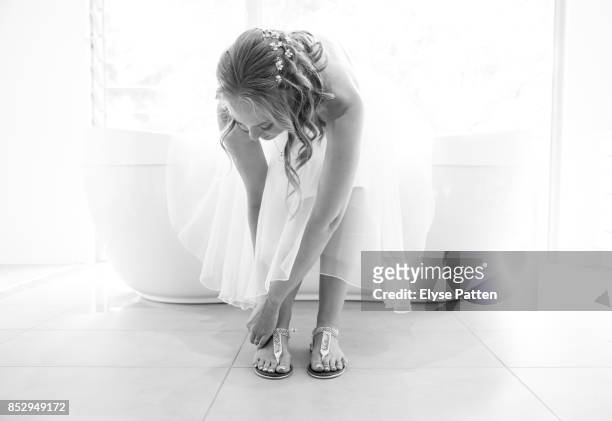 a bride is leaning over to buckle her casual shoes as the finishing touch to getting dressed for her wedding. she sits on a bathtub in front of a large window. - wedding shoes fotografías e imágenes de stock