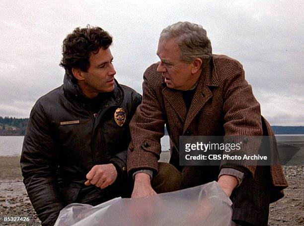Canadian actor Michael Ontkean and American actor Warren Frost talk as they examine a plastic-wrapped body on a rocky beach in a scene screen grab...