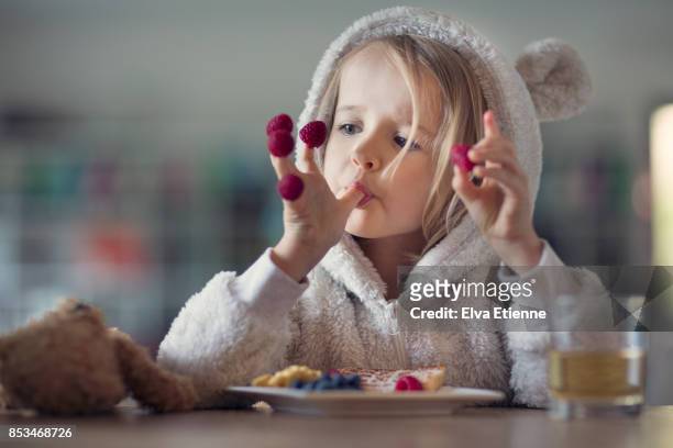 girl in cozy hooded pyjamas, eating raspberries off her fingers - fruta baya fotografías e imágenes de stock
