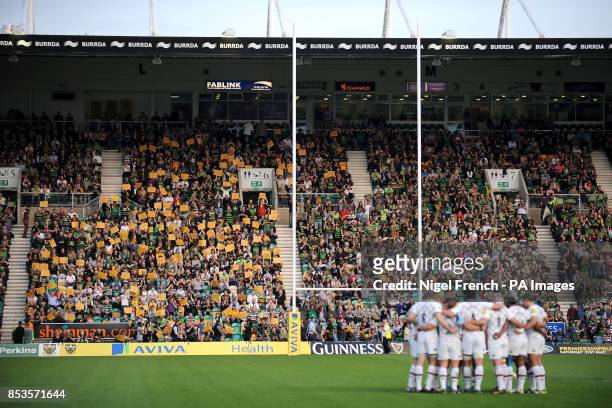 Northampton Saints fans hold up signs which read 'Come On You Saints' whilst the players gather in a huddle on the pitch