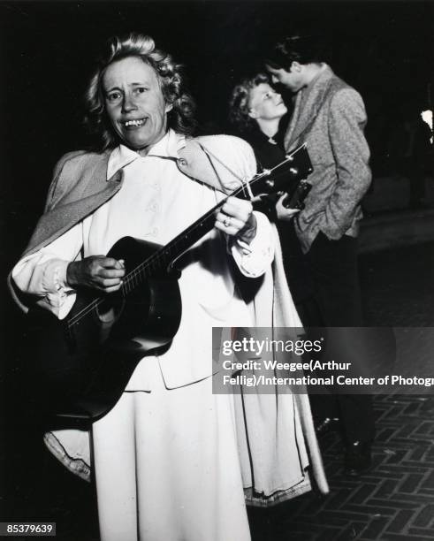 Woman plays the guitar on the sidewalk at night, and a man and woman stand in the background, New York, ca.1950s.