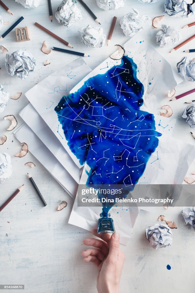 Crumpled paper balls with pencils and papers on a white wooden background with spilled ink forming starry sky with constellations. Writers hand holding an inkwell. Creative writing concept flat lay