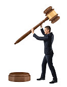 A small businessman on isolated white background holds and lowers a giant judge gavel on a sound block.