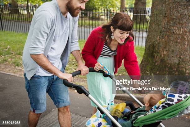 parents pushing pram, smiling at baby while walking to urban park. - baby pram in the park stock pictures, royalty-free photos & images