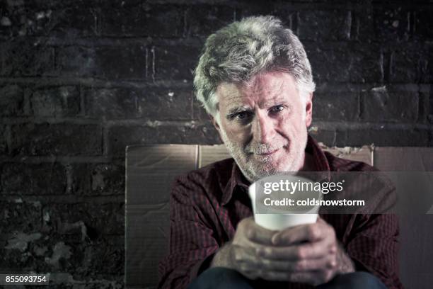 homeless senior male outdoors holding begging cup and looking at camera - hungry homeless stock pictures, royalty-free photos & images