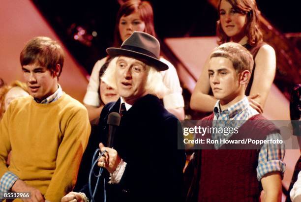 English DJ and television presenter Jimmy Savile stands with members of the studio audience on the set of a pop music television show in London circa...