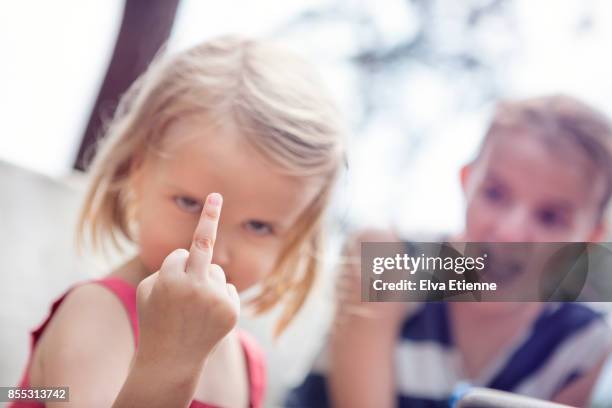 girl holding up a middle finger, with shocked sibling in the background - doigt dhonneur stockfoto's en -beelden