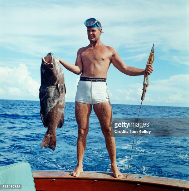 Portrait of American spearfisherman Art Pinder , a grouper in one hand and a fishing spear in the other, as he stands on the gunwale of a boat,...