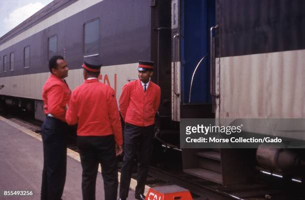 Scene on a station platform of three African American porters talking outside of a train car entrance, July, 1969. A footstool at the bottom of the...
