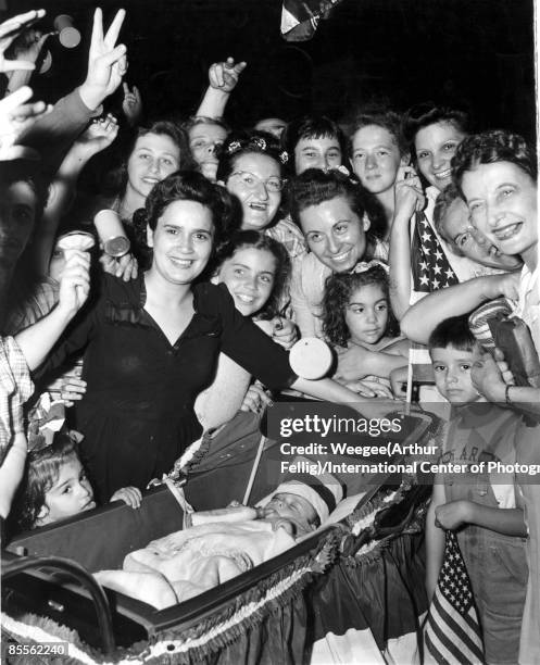 Crowd of people, some holding American flags and noise makers, with little Bobby De Marco, a baby aged 7 months in a carriage, celebrating the end of...