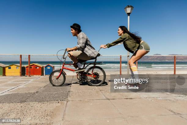 young man on bicycle towing girl on roller skates - offbeat stockfoto's en -beelden