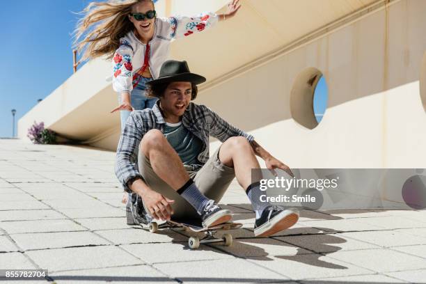 young woman pushing man sitting on skateboard - mens free skate imagens e fotografias de stock
