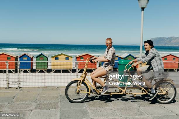 young couple riding a tandem bicycle on a boardwalk - bike beach stockfoto's en -beelden