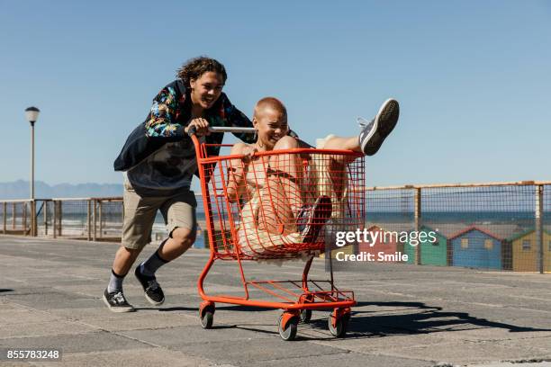 young man pushing girl in red trolley - friends smile stock-fotos und bilder