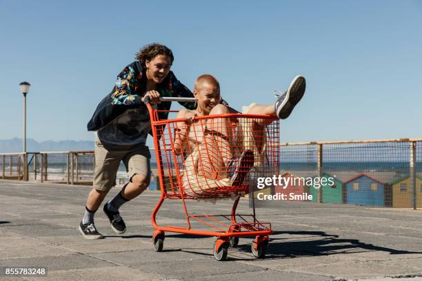 young man pushing girl in red trolley - young adults having fun stockfoto's en -beelden
