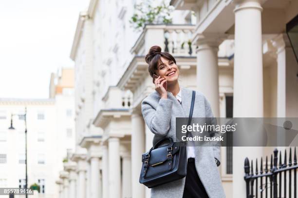 elegant beautiful woman talking on phone in front of city houses - gray coat stock pictures, royalty-free photos & images